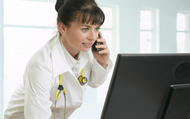 woman_doctor_stethoscope_around_neck_on_cordless_phone_looking_computer_telemedicine